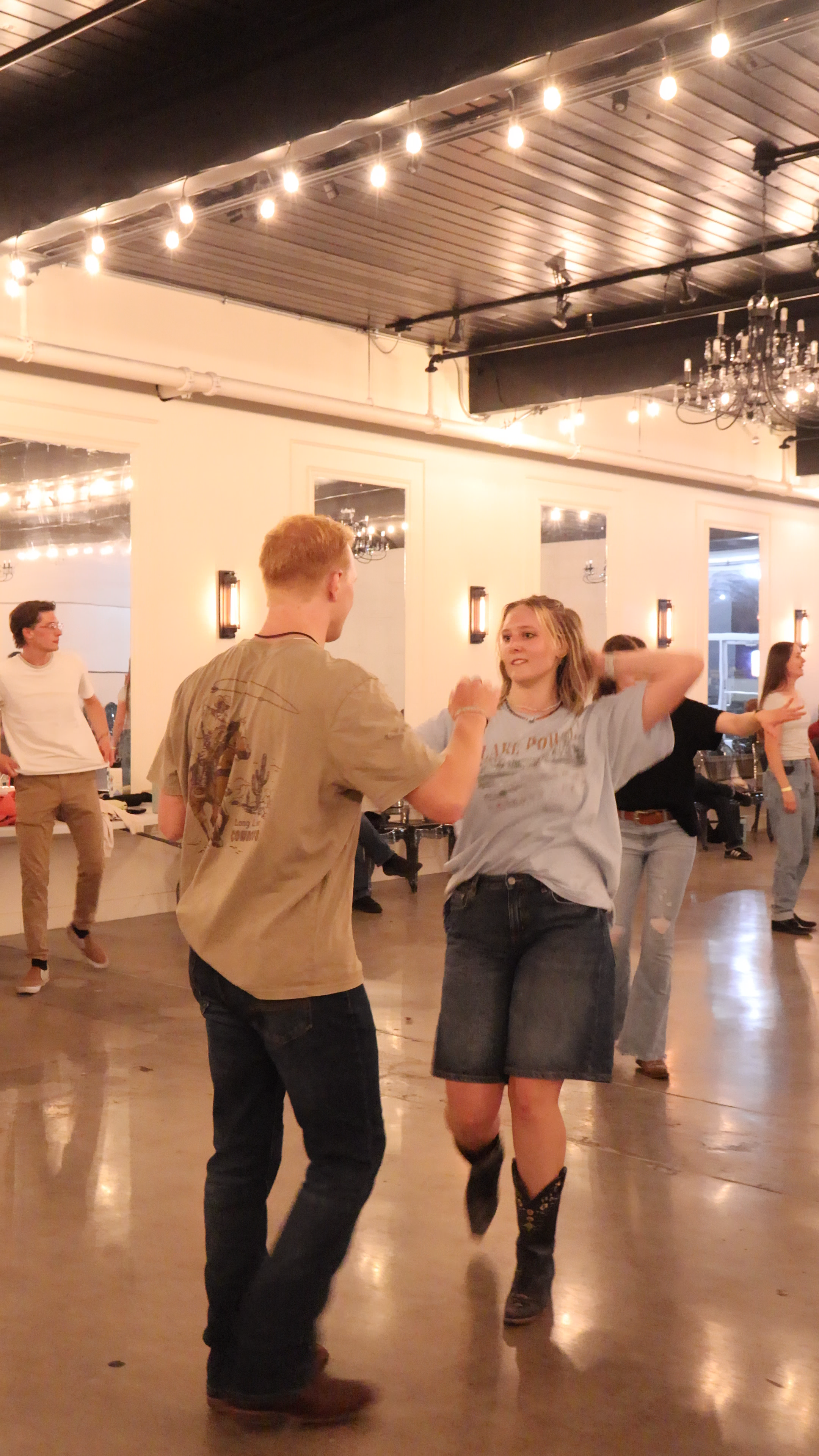 A group of young people country swing dancing together in a warmly lit room with string lights and mirrors, enjoying a social moment.