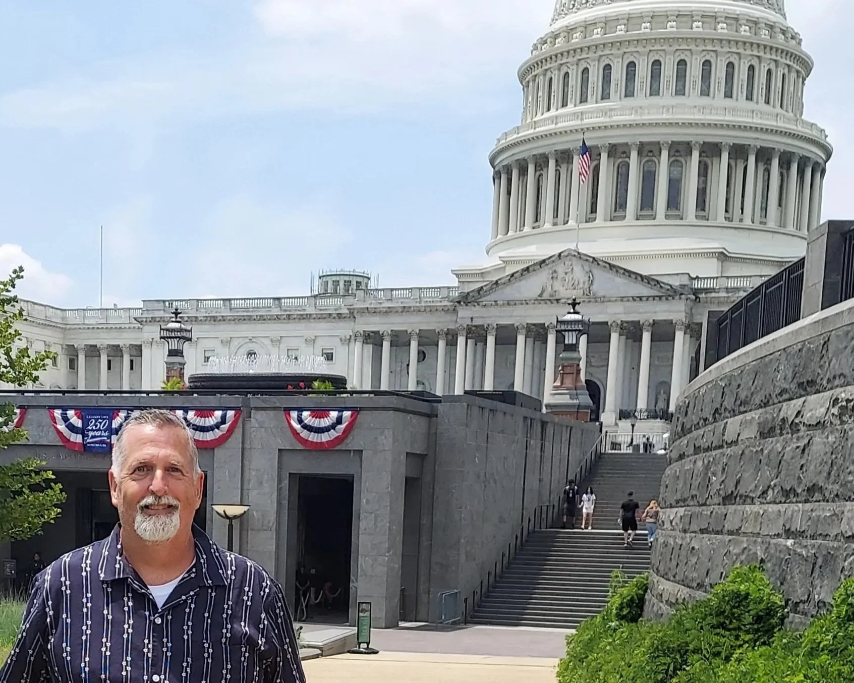 A smiling man with gray hair and a beard standing in front of the United States Capitol building decorated with patriotic banners.
