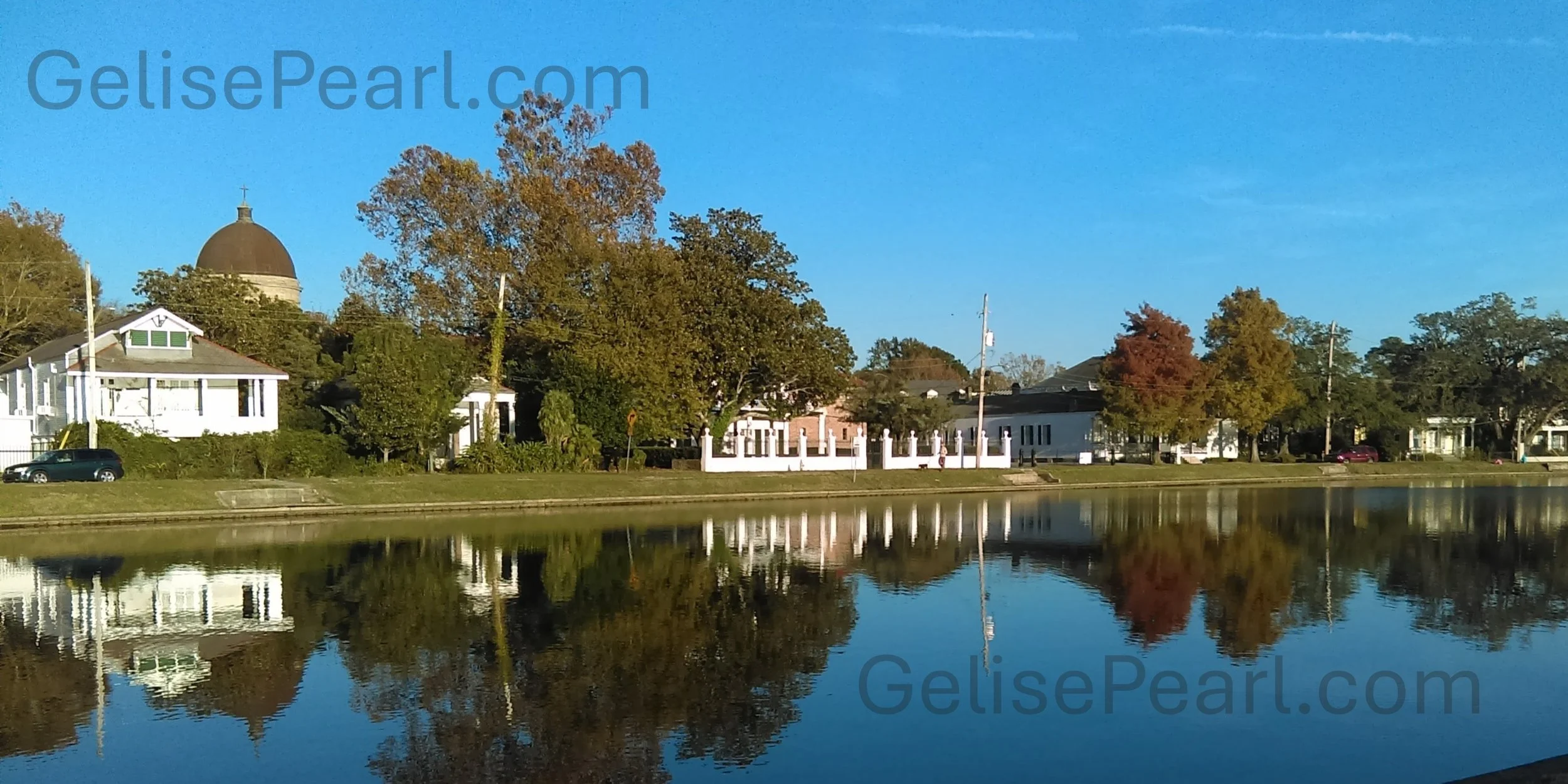 A scenic view of a calm river reflecting trees with green and orange leaves and houses on the opposite bank, under a clear blue sky at Bayou St. John in New Orleans