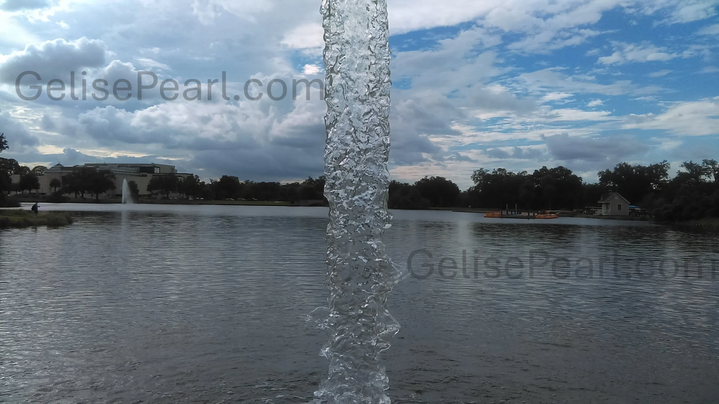 A water fountain splash with a lake and cloudy sky in the background, including trees and a building at the Big Lakes at City Park in New Orleans.