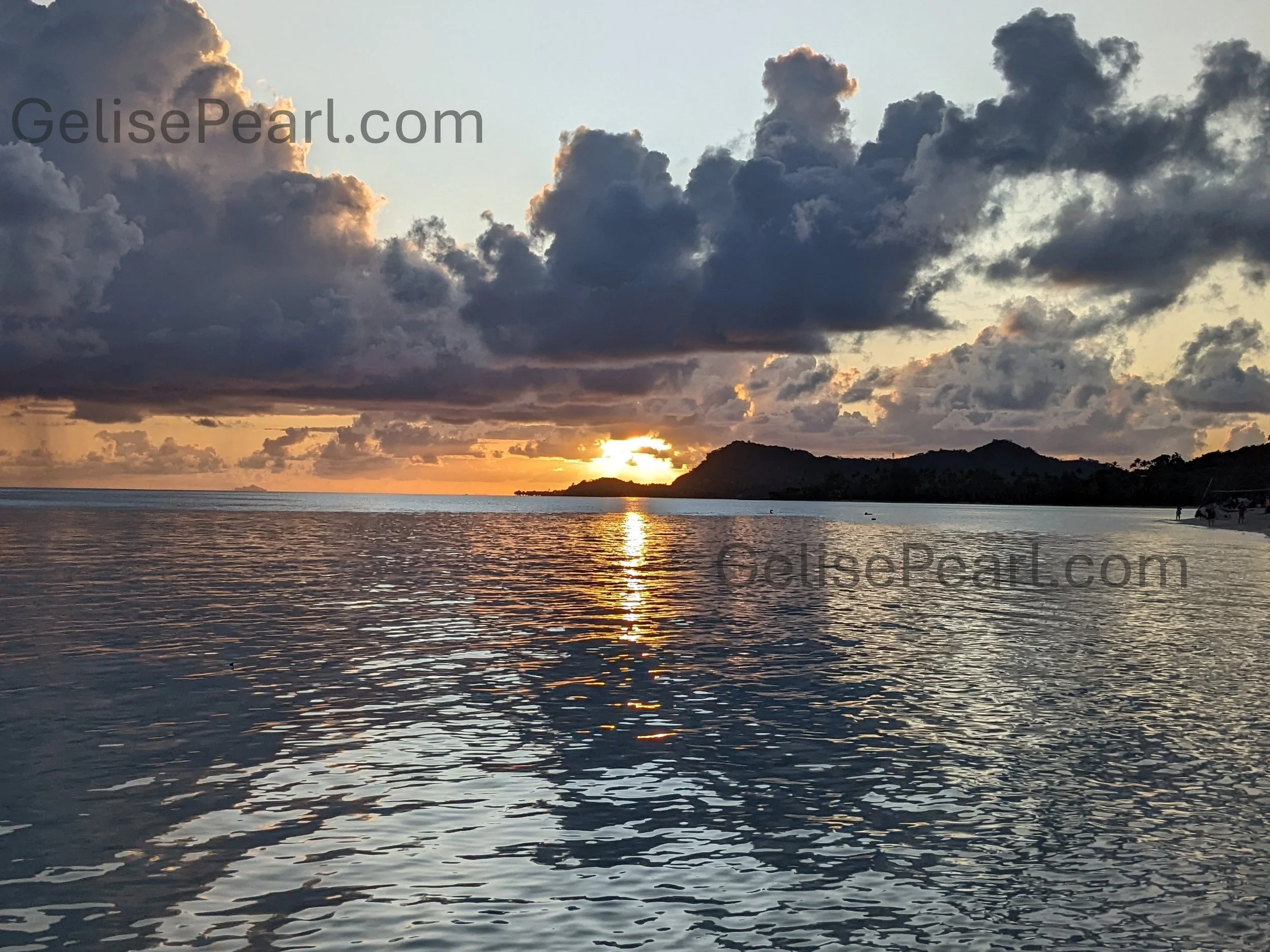 Sunset over the ocean with dark clouds in the sky and a silhouette of a hilly landscape in the distance.
