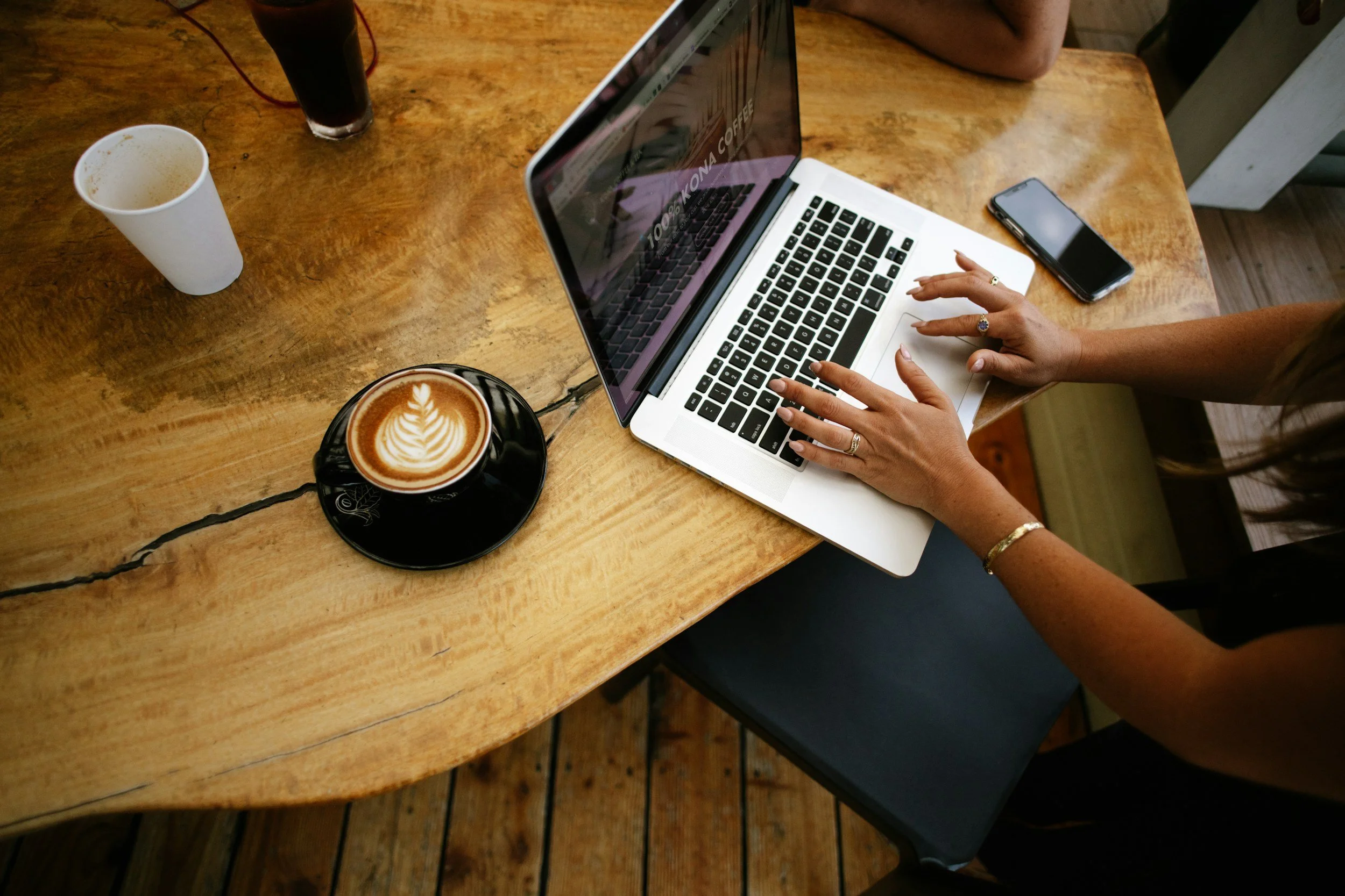 A person typing on a silver MacBook laptop at a wooden table in a coffee shop. There are cups of coffee, one with latte art, and a glass of dark soda on the table.