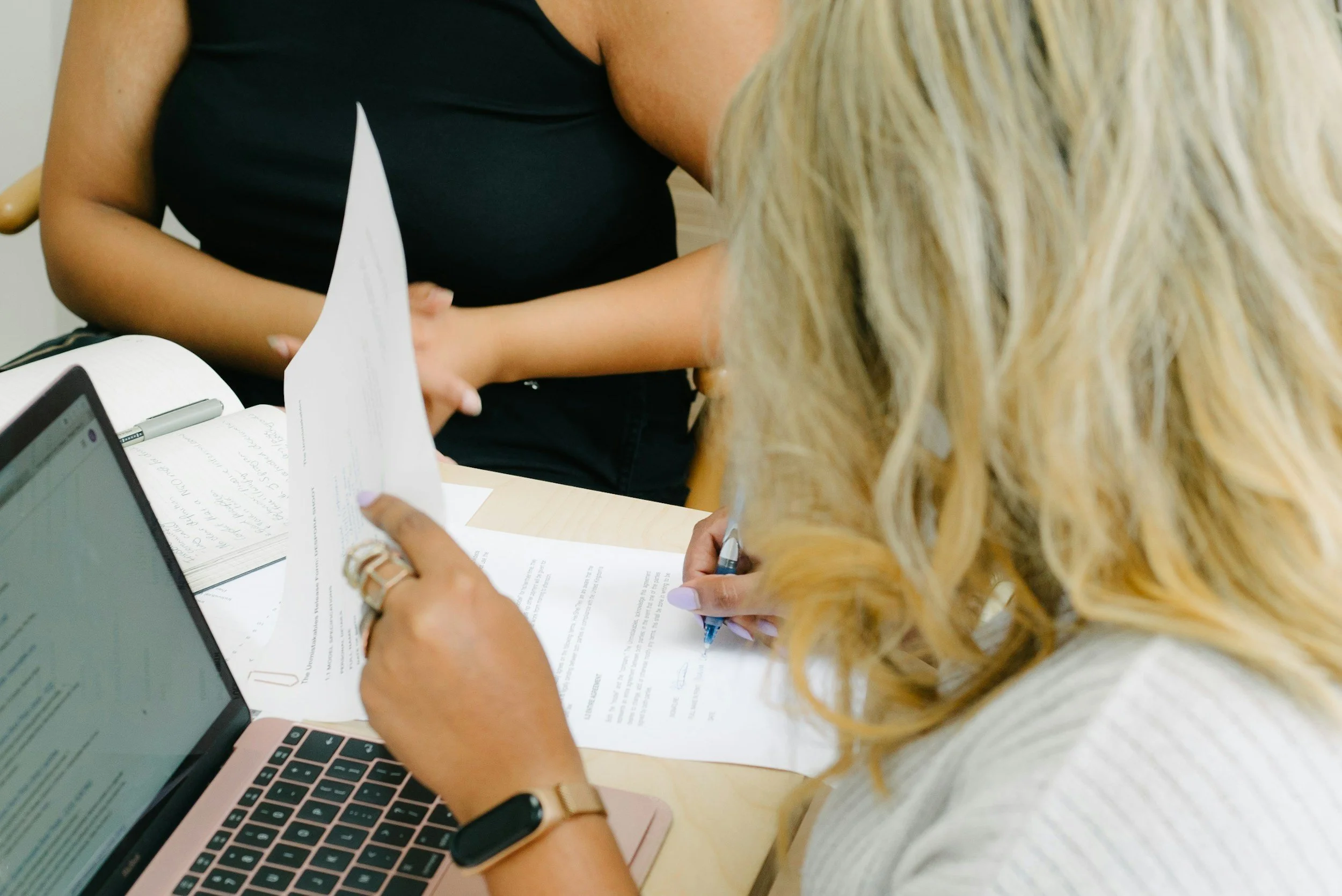 Two women sitting at a desk, one with long blonde hair writing on papers, and the other with medium brown skin and black top, holding a paper. A laptop and a notebook are also on the desk.