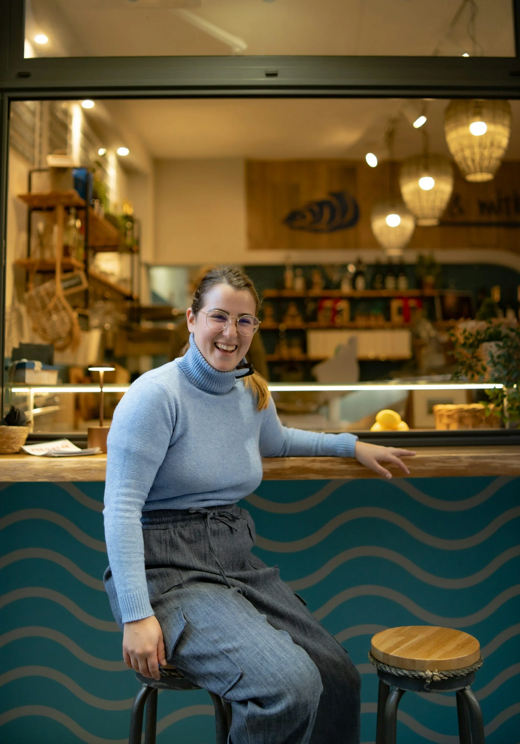 A woman with glasses and a blue sweater smiling and sitting on a stool inside a cafe, with a wooden counter and decorative lighting in the background.