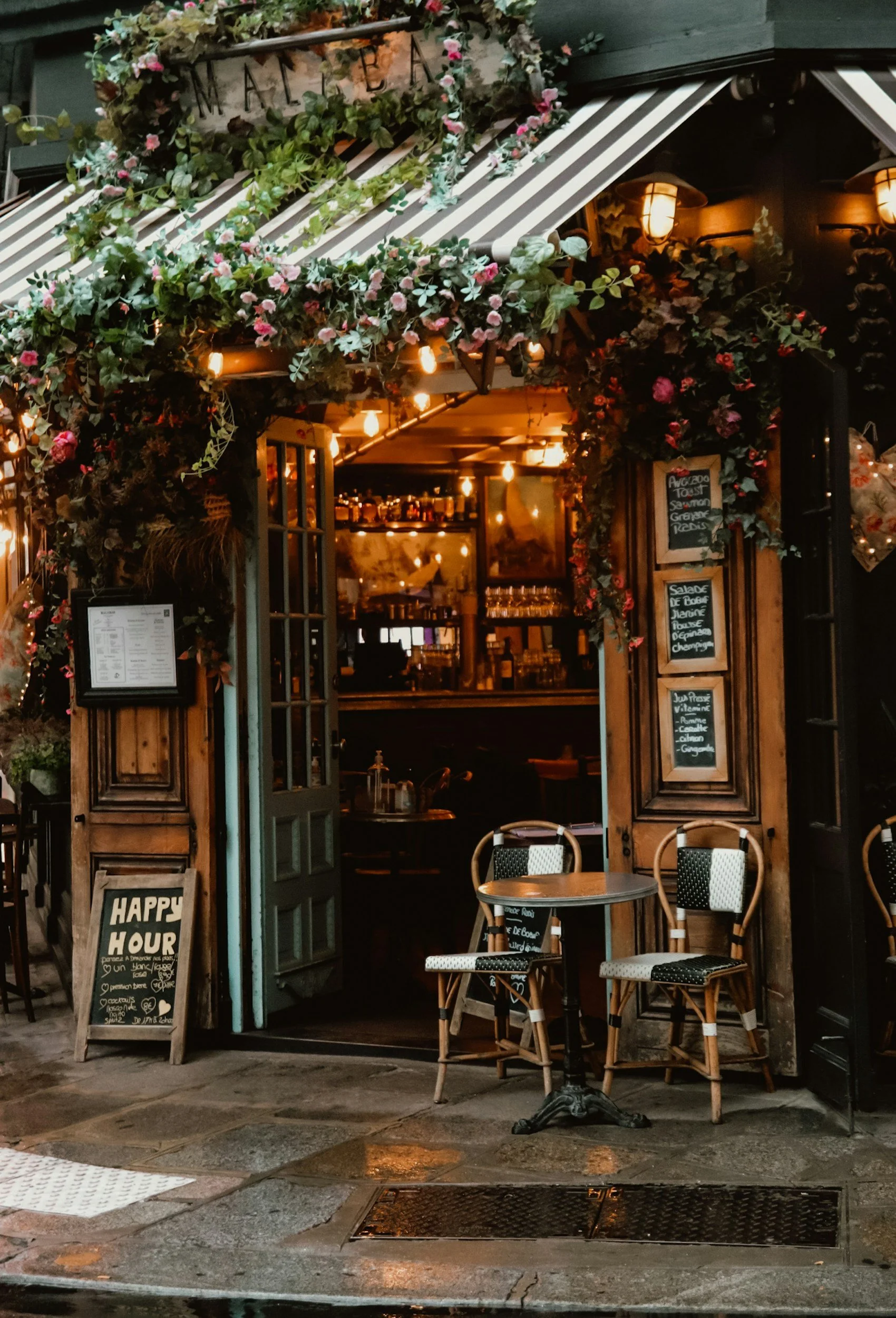 A cozy outdoor restaurant entrance decorated with greenery, flowers, and warm string lighting. There are a couple of small tables with chairs, a chalkboard menu, and a