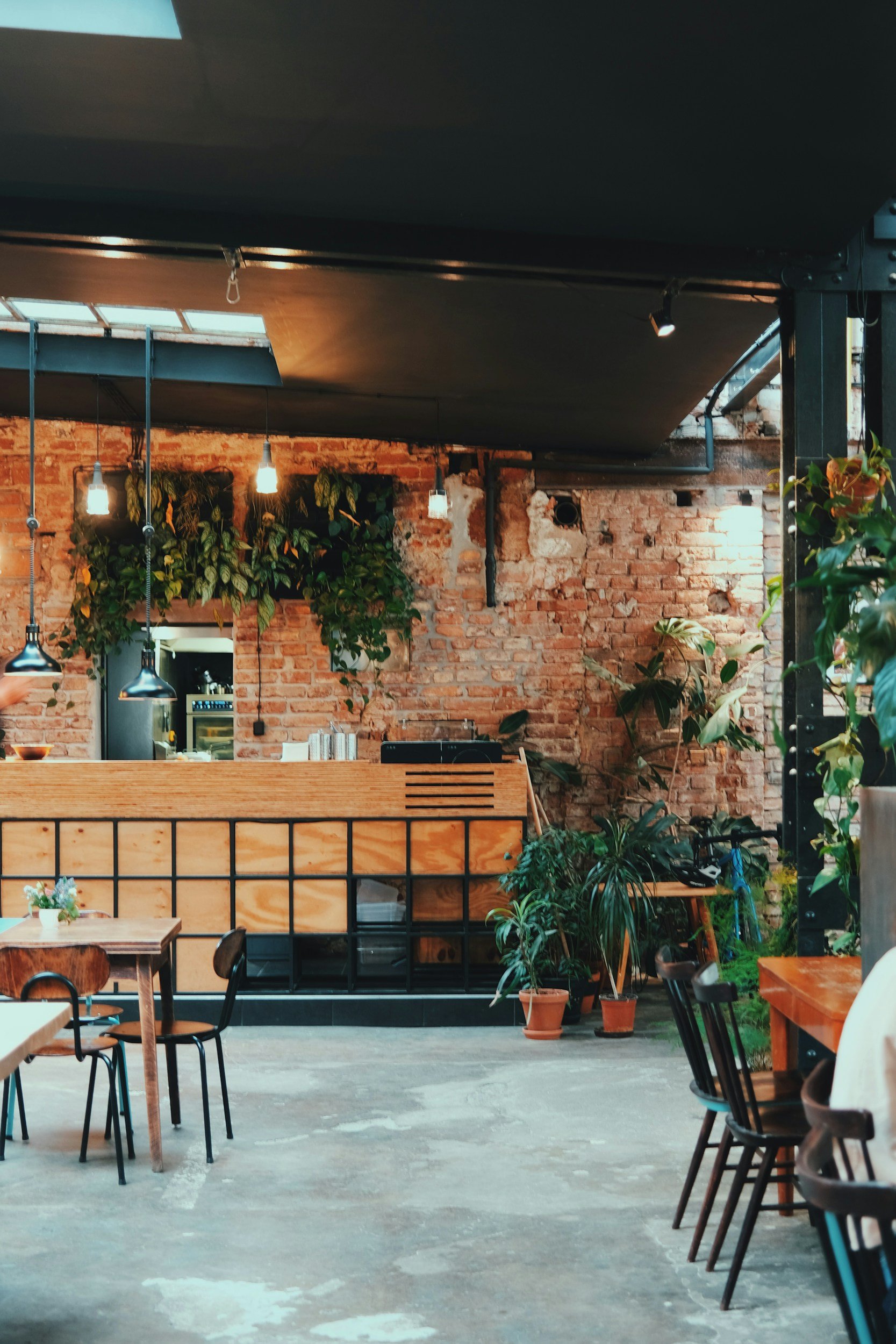 Interior of a modern restaurant or café with exposed brick walls, hanging light fixtures, and several tables and chairs, decorated with plants.