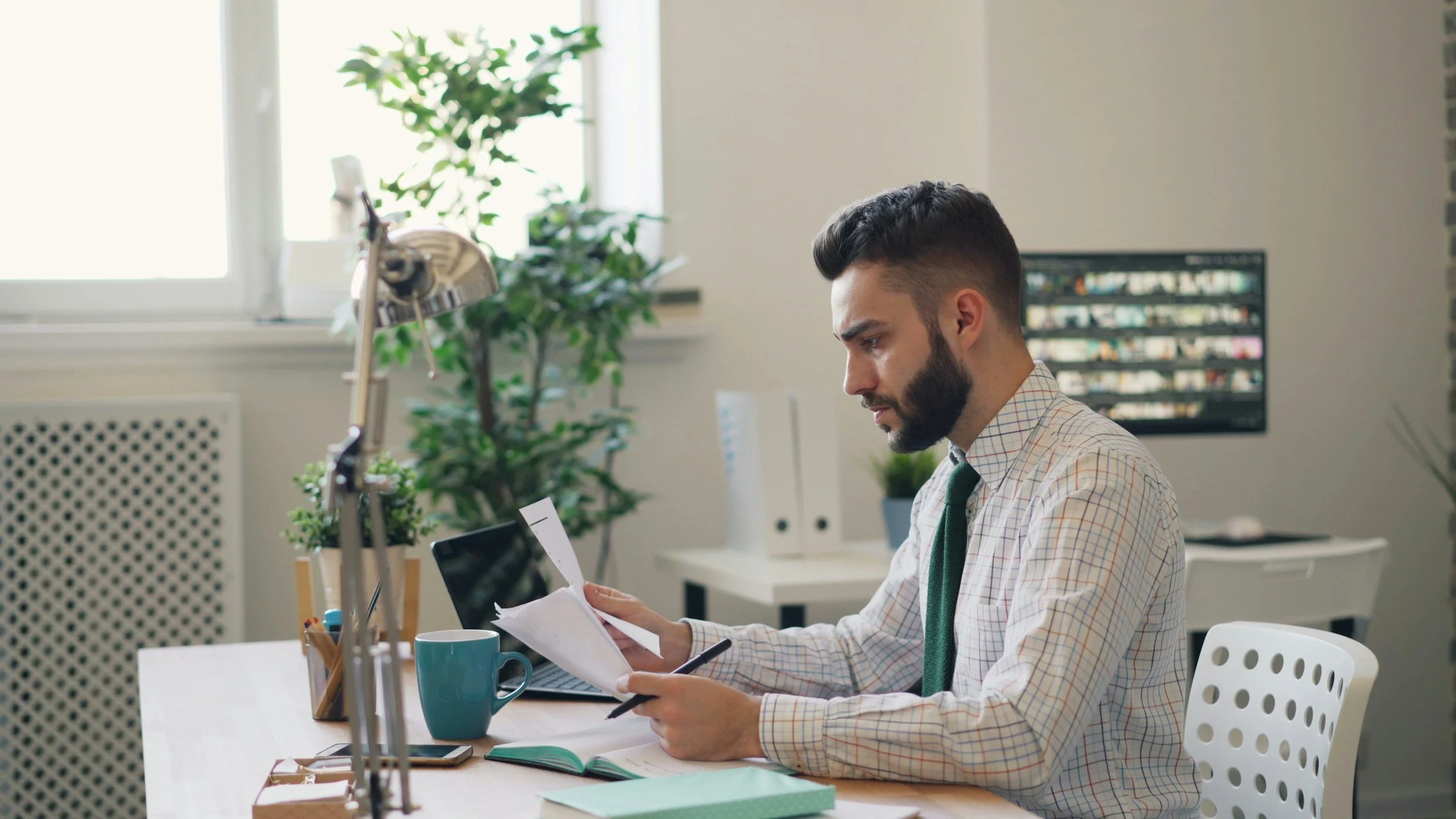 A man with dark hair and a beard, wearing a checked shirt and green tie, sitting at a desk in an office, reading a paper.