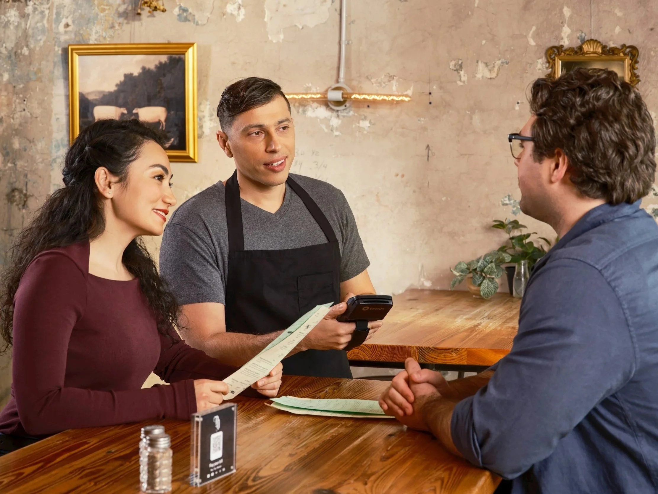 A woman and man facing each other at a restaurant table, with a waiter presenting a check. The woman holds a menu, and the man is listening to the waiter, who has a notepad and pen.