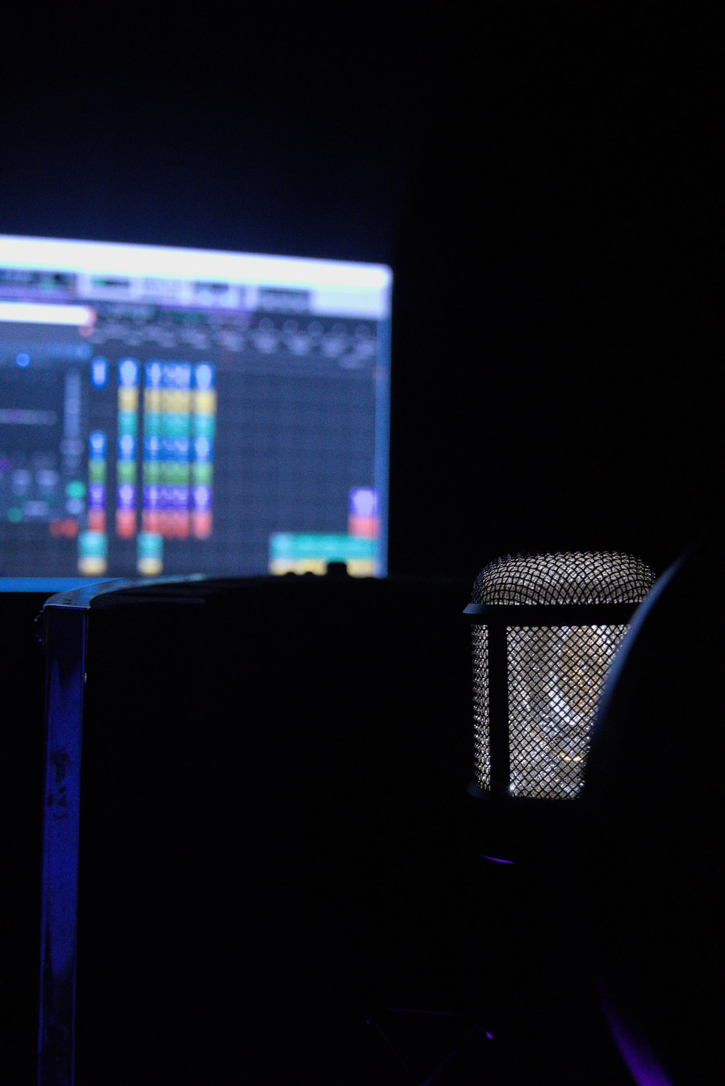 A computer screen displaying digital audio workstation software with colorful tracks and waveforms, with a black background, illuminated by blue and purple LED lights, and a mesh desk lamp in the foreground.