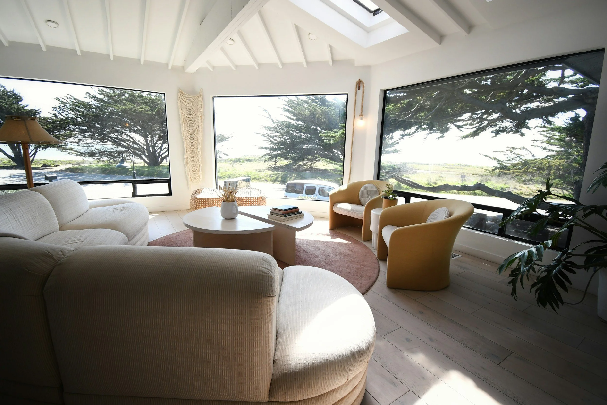 Living room with large windows showing trees outside, a white curved sofa, yellow armchairs, a round coffee table with books and a vase, and a ceiling skylight letting in natural light.