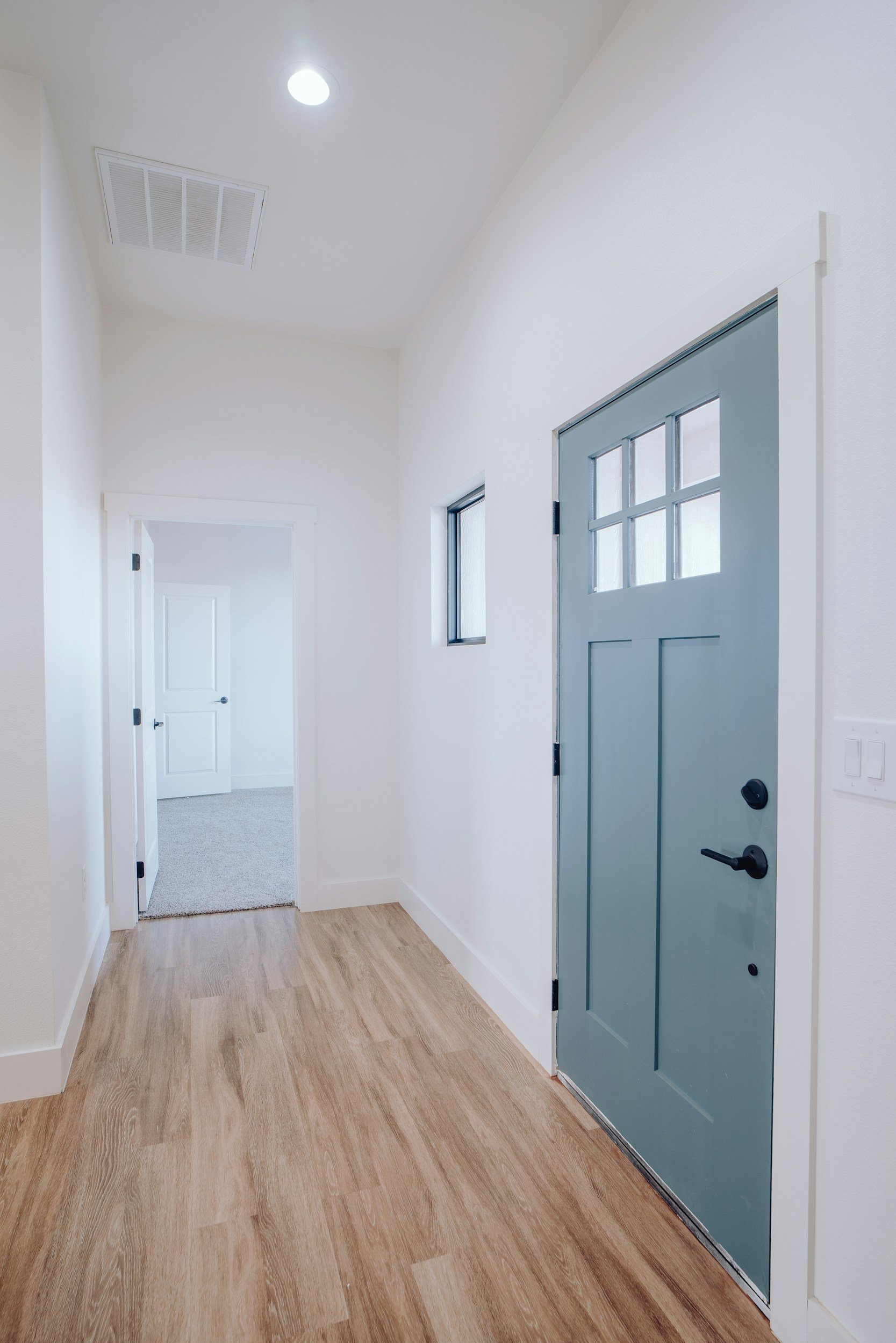 Interior view of a modern home entryway with light wood flooring, white walls, a blue front door with window panes, a small window beside it, and a ceiling vent and light fixture.