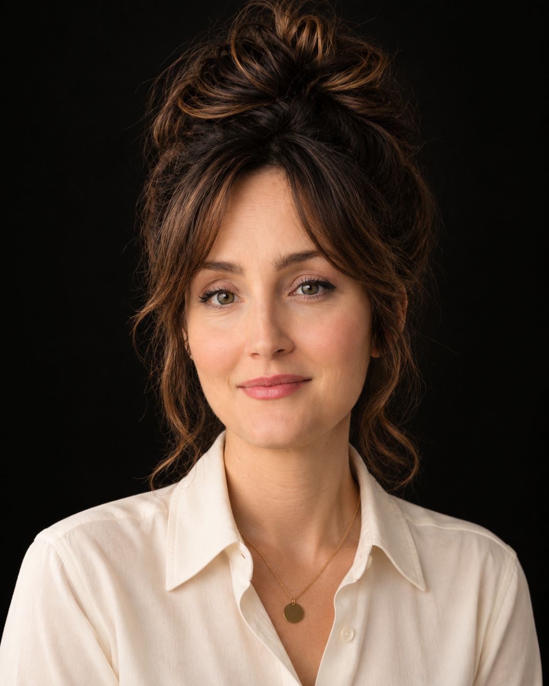 A woman with light skin and brown hair styled in a messy bun, wearing a cream-colored shirt and a gold necklace, smiling softly against a black background.
