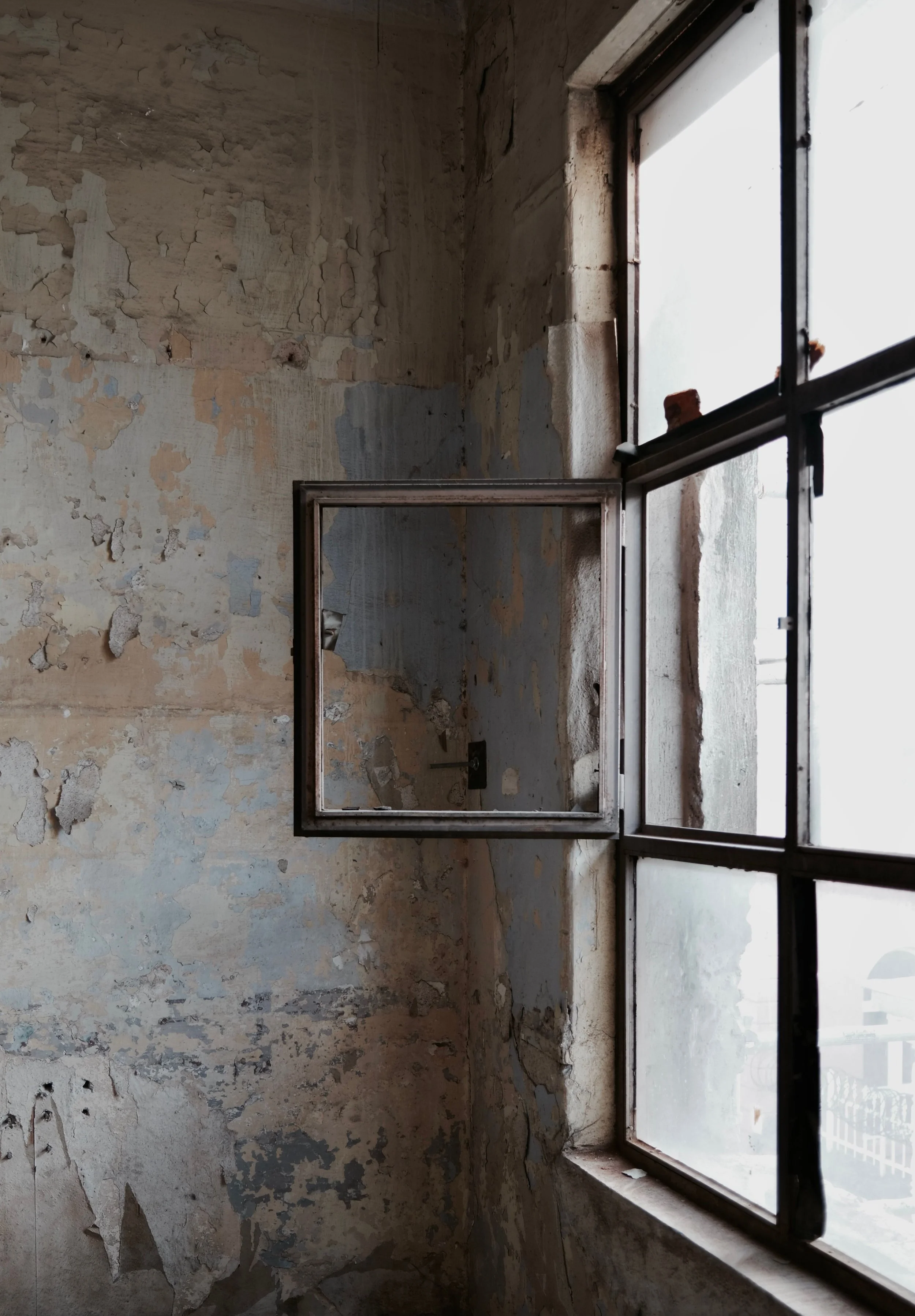 An old, dilapidated room with peeling paint on the walls and a large window with a metal frame, partially open. The window lets in light, illuminating the worn textures and cracks in the wall surfaces.