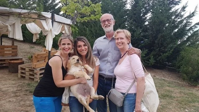 Quattro persone sorridenti, due donne e due uomini, con un cane, all'aperto in un giardino con sedie di legno e tende, in un momento conviviale.