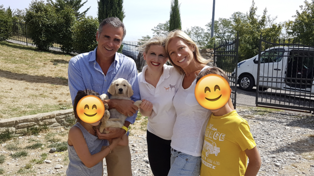 Quattro persone sorridenti con due bambini e un cane in un'area all'aperto, con un cancello nero e auto sullo sfondo.