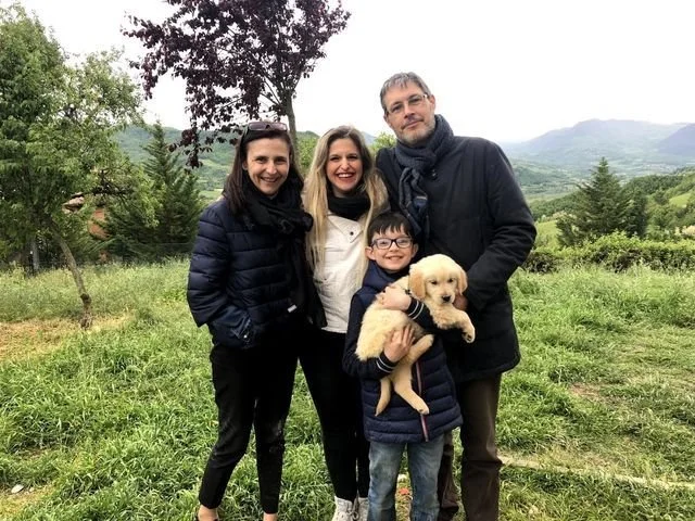 Quattro persone e un cane in un paesaggio naturale con alberi e montagne sullo sfondo.