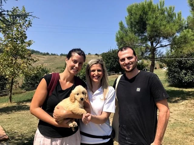 Tre persone sorridenti con un cucciolo di cane in un parco con alberi e cielo azzurro.