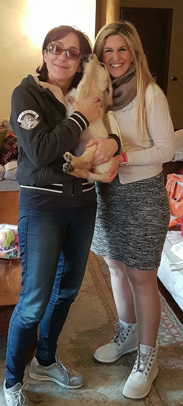 Due donne sorridenti tengono un cucciolo di cane tra di loro in un ambiente domestico.