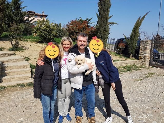 Quattro persone sorridenti e un cucciolo di cane in piedi all'aperto, con un'area naturale e un cancello sullo sfondo.