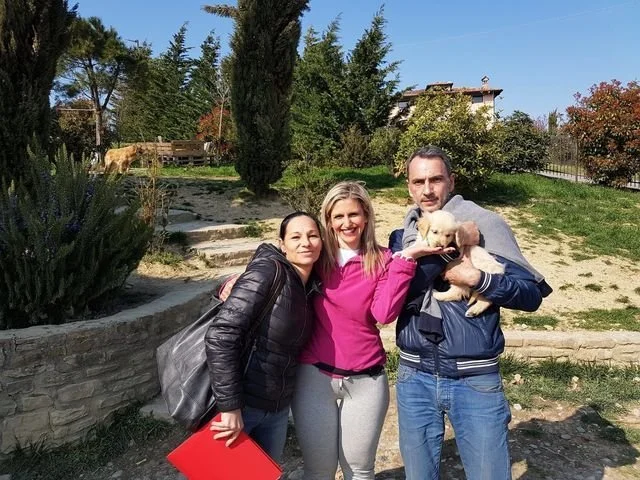 Tre persone sorridenti con un cane in un giardino soleggiato, con alberi e un edificio sullo sfondo.