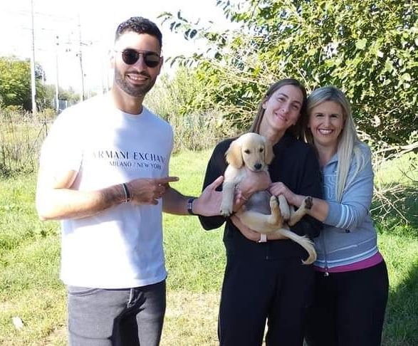 Tre persone sorridenti e una cucciola di cane in un'area verde soleggiata, con alberi sullo sfondo.