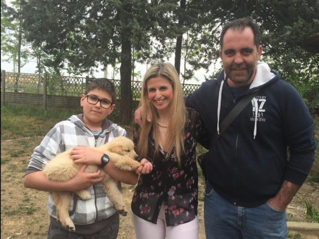 Tre persone sorridenti e un cane lupo giovane in un giardino con alberi e recinzione in sfondo.