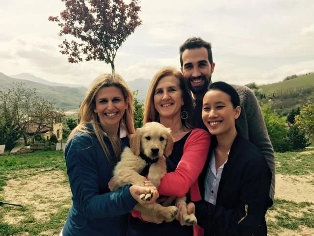 Quattro persone sorridenti e un cucciolo di cane in un paesaggio naturale con alberi e montagne sullo sfondo.