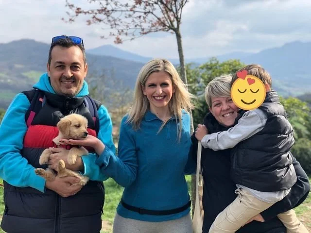 Gruppo di quattro persone con un cane, sorridenti, in un paesaggio naturale con montagne e alberi sullo sfondo.