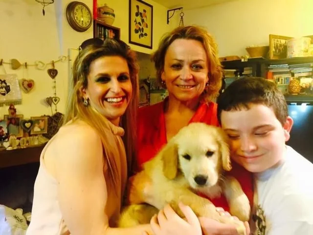 Tre persone sorridenti e un bambino con un cucciolo di cagnolino in casa, sfondo con decorazioni e scaffali.