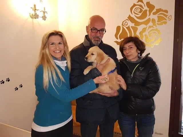 Tre persone con un cucciolo di cane in un ambiente interno, decorato con un muro con rose dorate e adesivi di zampette di cane.