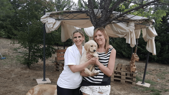 Due donne sorridenti che tengono un cucciolo di cane, sotto un gazebo in giardino con alberi in background.