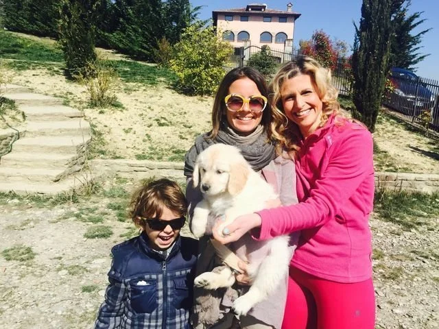Due donne sorridenti, una con occhiali da sole, tengono un cucciolo di cane bianco, insieme a un bambino con occhiali scuri, in un'area all'aperto con vegetazione e una casa sullo sfondo.