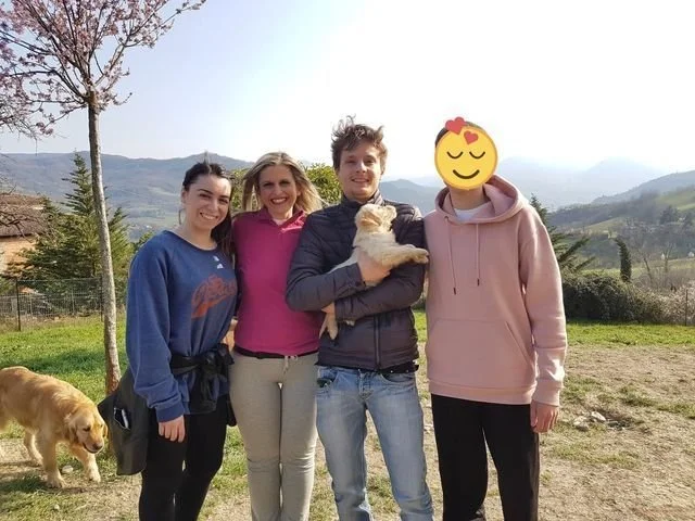 Quattro persone e due cani in un campo con paesaggi collinari sullo sfondo. Due donne e due uomini, uno degli uomini tiene un cane in braccio. Una donna ha il volto coperto da un'emoji.