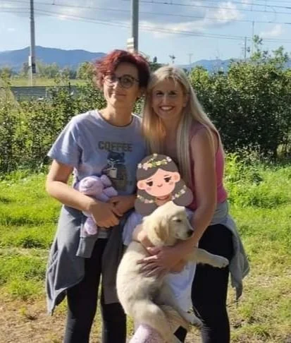 Due donne sorridenti che abbracciano un cucciolo di cane in un'area verde all'aperto con alberi e montagne sullo sfondo.
