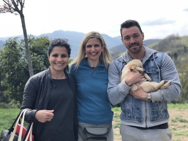 Tre persone sorridenti che posano all'aperto con un cane, con paesaggi collinari sullo sfondo.