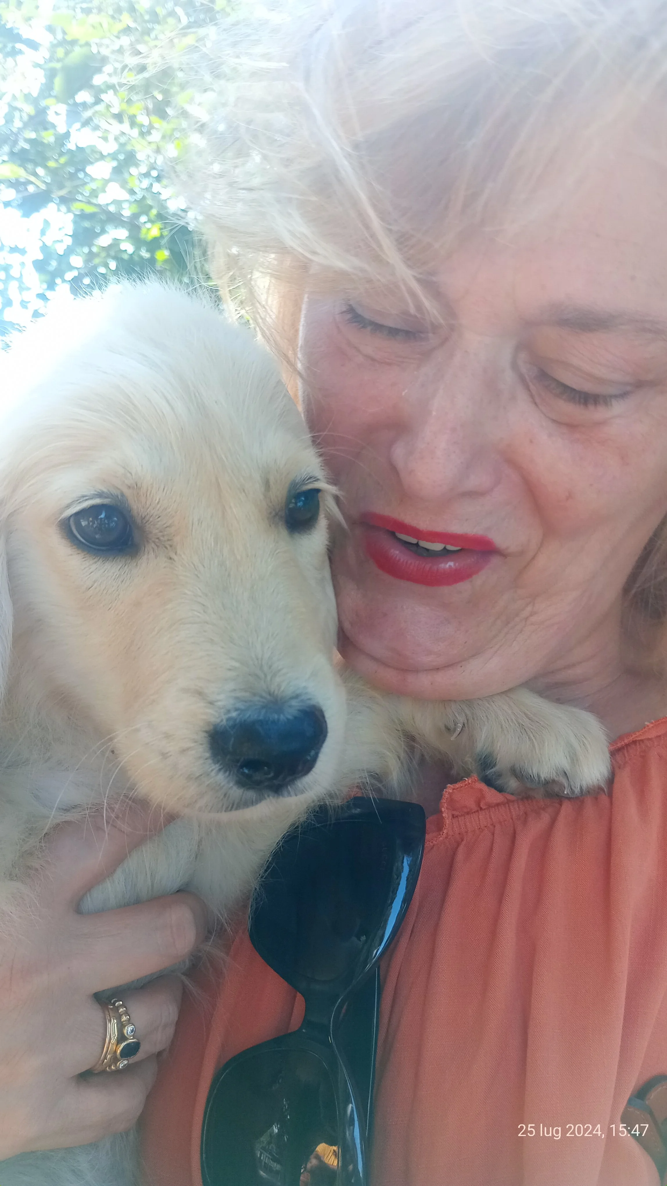 Una donna con i capelli biondi e rossetto rosso tiene in braccio un cucciolo di cane bianco con occhi azzurri. C'è luce naturale e alberi sullo sfondo.