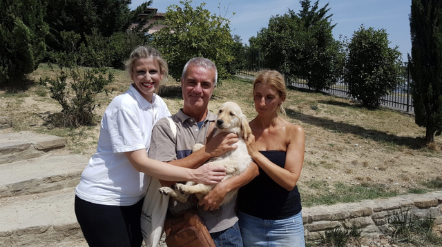 Tre persone e un cane in un parco, sorridenti, con alberi e cespugli sullo sfondo.
