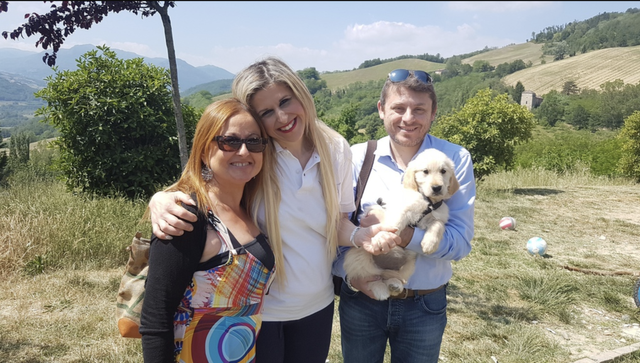 Tre persone sorridenti in un campo aperto con un cane, circondate da colline e foreste.