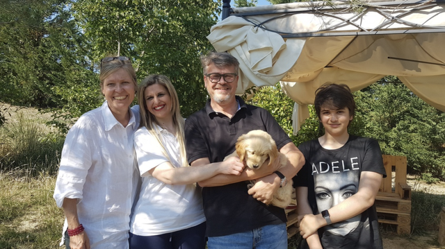 Quattro persone sorridenti e un cucciolo di cane in un contesto all'aperto con alberi e una tenda