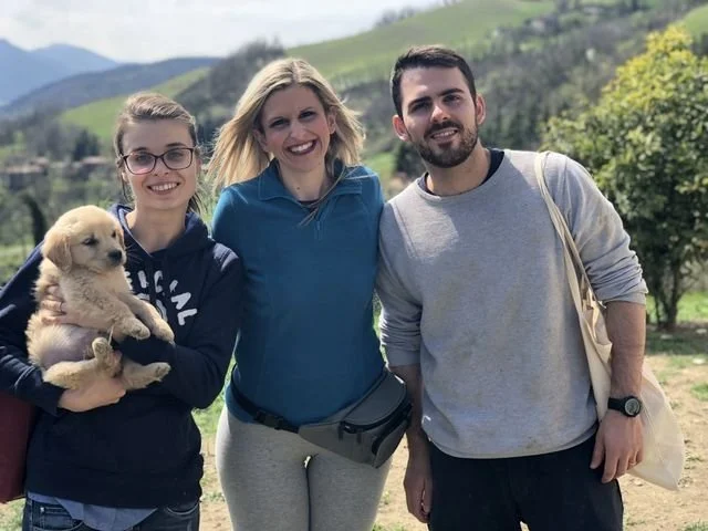 Tre persone sorridenti e una cucciola di cane in piedi in una campagna con colline verdi sullo sfondo.
