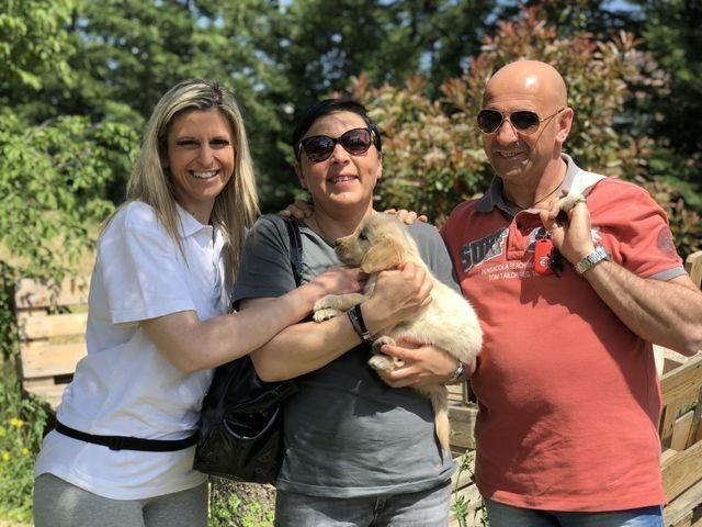 Tre persone sorridenti con un cucciolo di cane in un giardino soleggiato.