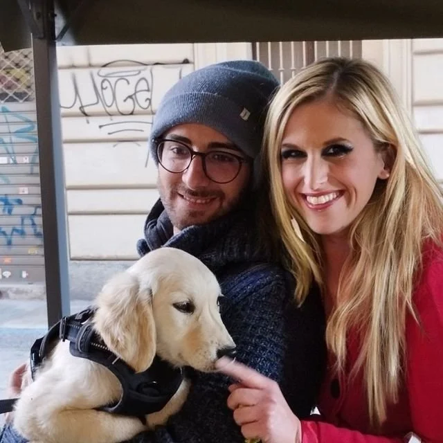 Una coppia sorridente con un cane da guida di razza Golden Retriever, seduti in un'area esterna.