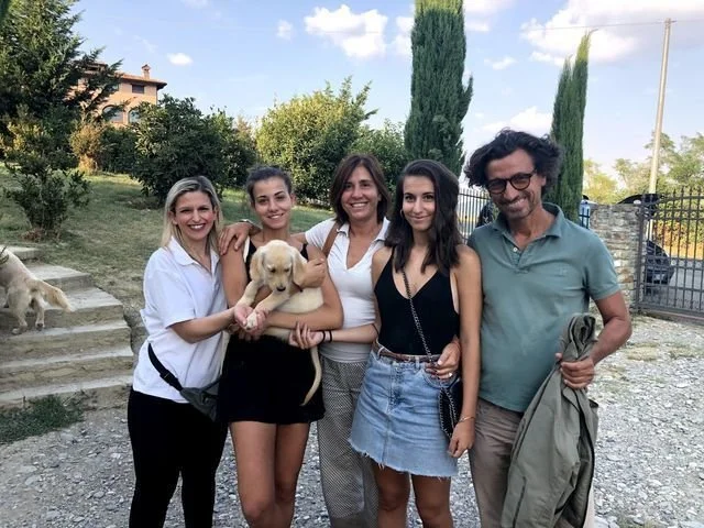 Cinque persone sorridenti con un cane giallo in un'area aperta con alberi e un cancello.