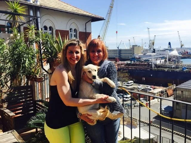 Due donne sorridenti con un cane bianco in un'area all'aperto vicino al porto con navi e gru sullo sfondo.