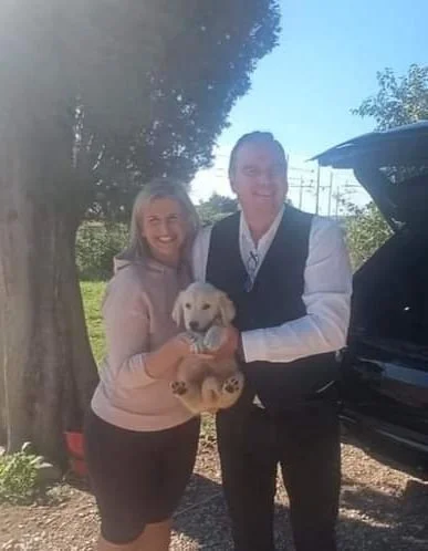 Due persone sorridenti tengono un cucciolo di cane tra di loro, in un ambiente all'aperto con alberi e un'auto sullo sfondo.