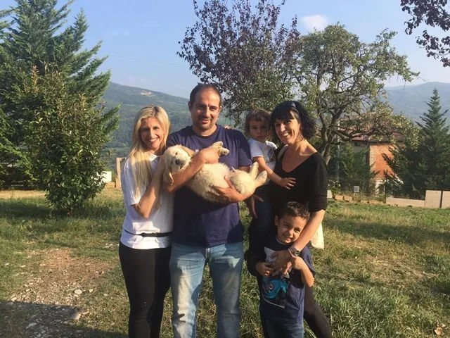 Una famiglia di cinque persone con un cane in un giardino con alberi e montagne sullo sfondo.