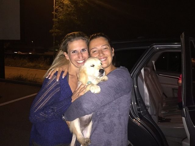 Due donne sorridenti che abbracciano un cane di razza husky criollo in un parcheggio notturno.