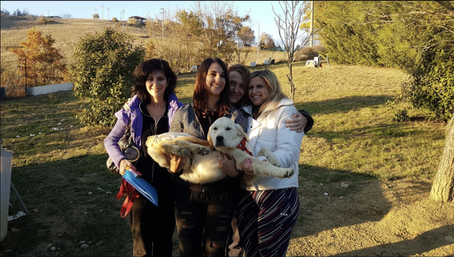 Quattro donne sorridenti che tengono un cucciolo di cane in un'area verde all'aperto durante l'autunno, con alberi e colline sullo sfondo.