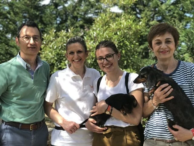 Quattro persone sorridenti con due cuccioli di cane, all'aperto con sfondo di alberi verdi.