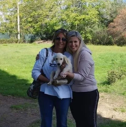 Due donne sorridenti che tengono un cucciolo di cane in un parco verde con alberi e cielo azzurro.